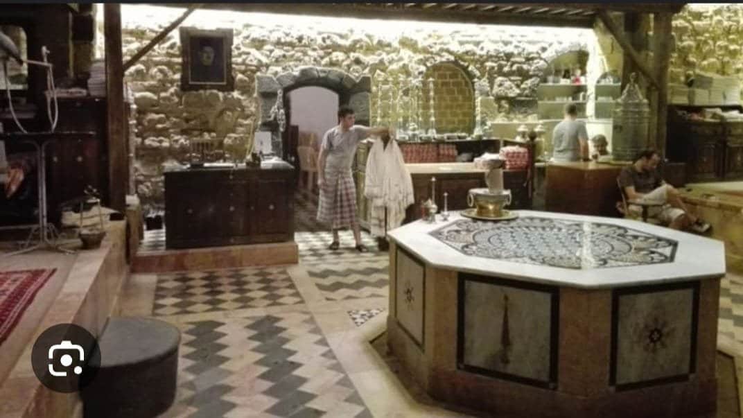 A view of a traditional Damascus public bathhouse, featuring ornate tile patterns, a central fountain, and visitors enjoying the space.